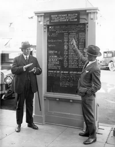 two men standing in front of a chart of ship arrivals circa 1930